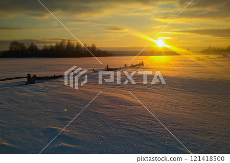 Wonderful winter landscape during an amazing sunset with a country road, passing near the snow-covered fence in full growth. In the background are seen thickets of wild willow 121418500