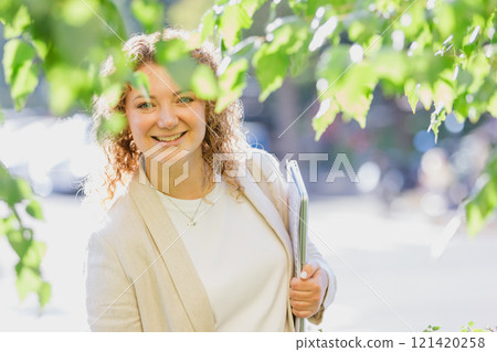 Professional woman is standing outdoors amidst lush greenery, holding documents 121420258