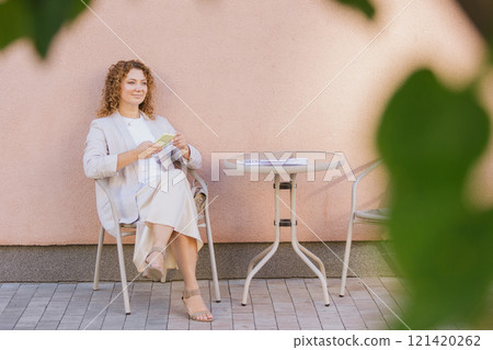 Woman in business attire sits at an outdoor cafe table, engrossed in her smartphone 121420262