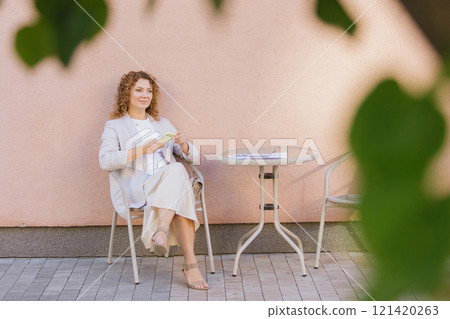 Woman in business attire sits at an outdoor cafe table, engrossed in her smartphone 121420263