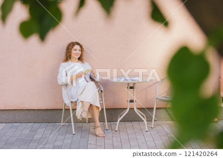 Woman in business attire sits at an outdoor cafe table, engrossed in her smartphone 121420264