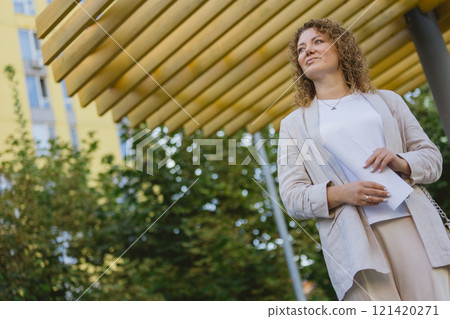 Businesswoman stands outdoors in an urban setting, holding documents and looking confident Businesswoman stands outdoors in an urban setting, holding documents and looking confident 121420271