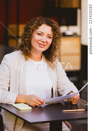 A businesswoman with curly hair is seated at an outdoor cafe, intently reading a document. A businesswoman with curly hair is seated at an outdoor cafe, intently reading a document. 121420283