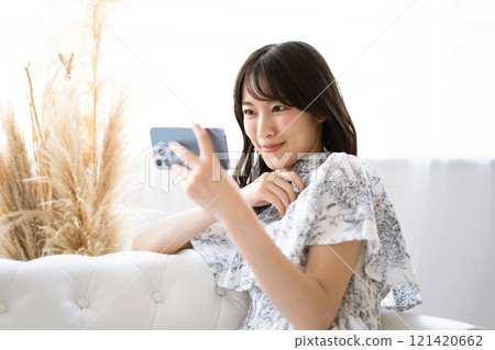 長黑髮的年輕女子坐在白色沙發上，手上拿著智慧型手機 121420662
