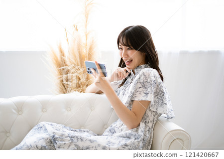 A young woman with long black hair sitting on a white sofa and holding a smartphone A young woman with long black hair sitting on a white sofa and holding a smartphone 121420667