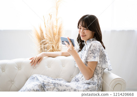A young woman with long black hair sitting on a white sofa and holding a smartphone A young woman with long black hair sitting on a white sofa and holding a smartphone 121420668