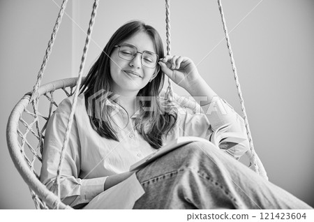 Portrait of girl relaxing with book on indoor swing with closed eyes. Pretty female in glasses excited about story she reading. Cozy afternoon with book in hanging chair in home. Black and white image 121423604
