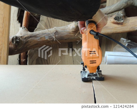 view of orange electric jigsaw cutting large piece of thick cardboard or plywood, cutting line of compressed wood material with jigsaw 121424284