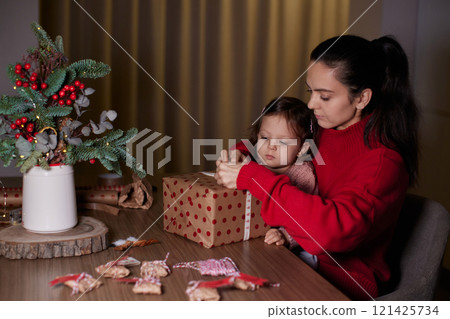 mother and her little child girl together wrapping Christmas gifts at home in the evening, preparing presents for family. 121425734
