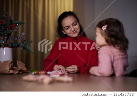 mother and her little child girl together wrapping Christmas gifts at home in the evening, preparing presents for family. 121425738