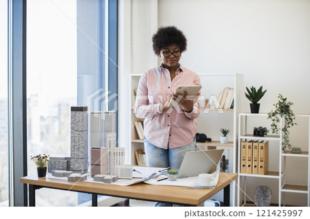 African American female architect in modern office designs city project using digital tablet. She stands by table with scale models, focused on innovation and creativity. African American female architect in modern office designs city project using digital tablet. She stands by table with scale models, focused on innovation and creativity. 121425997