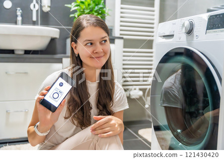 Young woman using mobile phone control via wi-fi and app for washing machine in home. Homesmart and new technology concept Young woman using mobile phone control via wi-fi and app for washing machine in home. Homesmart and new technology concept 121430420