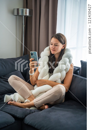 Young woman sits on a sofa with a weighted pillow on her shoulders and does a therapeutic practice aimed at getting rid of feelings of loneliness, session with a neurocoach 121430427