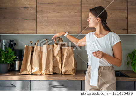 Young woman with receipt from the store next to paper bags full of food in kitchen. Groceries ordered online and delivered by courier 121430429