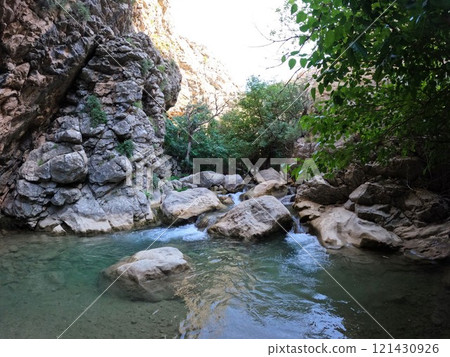 Mountain river with clear cold water with large stones against the background of rocks, trees and bushes, sunlight flooding the rocks in the background Mountain river with clear cold water with large stones against the background of rocks, trees and bushes, sunlight flooding the rocks in the background 121430926
