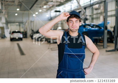 Confused car mechanic male in overalls feeling puzzled holding visor of his cap, looking at camera with serious expression. Technician man doing car maintenance in auto repair workshop. Confused car mechanic male in overalls feeling puzzled holding visor of his cap, looking at camera with serious expression. Technician man doing car maintenance in auto repair workshop. 121433169