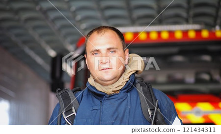 Portrait of tired male firefighter standing near a fire engine at department. Young exhausted fireman in protective uniform looking at camera after fire against the background of big red truck 121434311