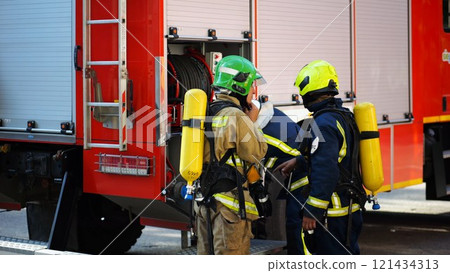 Young firemen prepare to extinguishing fire near big truck. Male firefighters in full equipment ready to putting out fire near a red car. Concept of saving lives and heroic profession. Slow motion 121434313