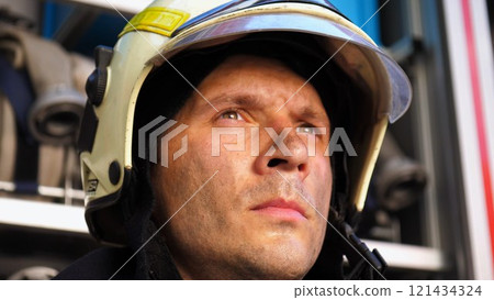 Young tired fireman in protective uniform against the background of a fire engine. Portrait of male firefighter with dirty face near fire truck. Concept of saving lives and heroic profession. Close up 121434324
