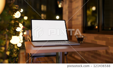 A close-up of a laptop on a table on an apartment balcony at night, illuminated by bulk lights. 121436588