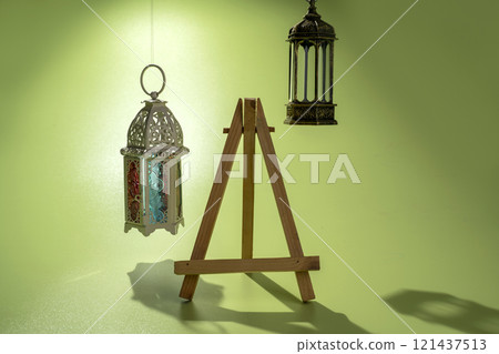 Wooden easel and Arabic lanterns hanging on a colored background. Decoration for Muslim holidays celebration 121437513