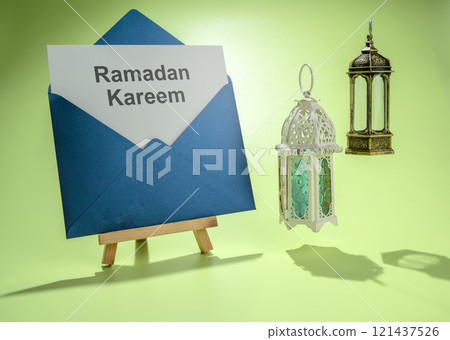 Wooden easel and envelope with text of Ramadan Kareem and Arabic lanterns hanging on a colored background 121437526