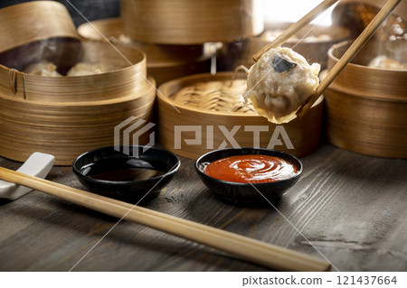 Hand with chopstick eating Chinese dim sum dumplings on bamboo wooden basket bowl with sauce and soy sauce on wooden table 121437664
