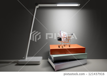 A white desk lamp illuminated a dollar bill on a shopping cart on a stack of books on a white table against a dark background A white desk lamp illuminated a dollar bill on a shopping cart on a stack of books on a white table against a dark background 121437916