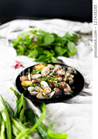 Garlic clams and mussels are presented on a black plate, surrounded by green herbs and chili. 121438285