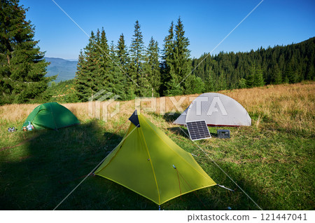 Tourist tent with solar panel and portable power station nearby in grassy field. Scene surrounded by rolling hills, tall pine trees under clear blue sky, showcasing eco-friendly camping setup. 121447041
