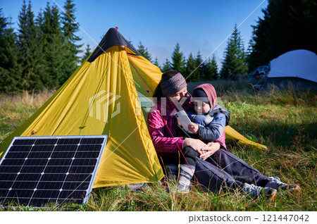 Young child and mother play with smartphone while charging with photovoltaic solar panel near tourist tent in summer. Integration of renewable energy in outdoor camping activities. Young child and mother play with smartphone while charging with photovoltaic solar panel near tourist tent in summer. Integration of renewable energy in outdoor camping activities. 121447042
