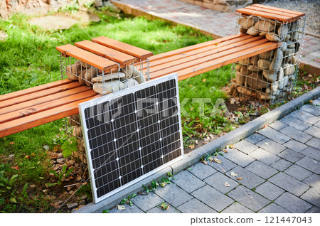 Photovoltaic solar panel solar panel against bench situated on paved walkway. Green grass and surrounding foliage highlight integration of sustainable renewable energy sources within city environments 121447043