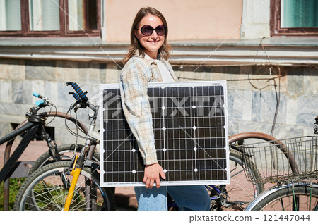 Happy woman holding solar panel, standing beside several parked bikes. Happy female wearing sunglasses and plaid shirt, enjoying sunny day in urban setting. 121447044