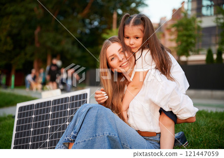 Mother and daughter sit together on grass, embracing in warm moment with solar panel beside in the evening. Concept of renewable energy and family, highlighting commitment to sustainable future. Mother and daughter sit together on grass, embracing in warm moment with solar panel beside in the evening. Concept of renewable energy and family, highlighting commitment to sustainable future. 121447059