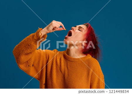 Woman taking yellow pill, symbolizing self-treatment to manage viral infection symptoms or fever against blue studio background. 121449834