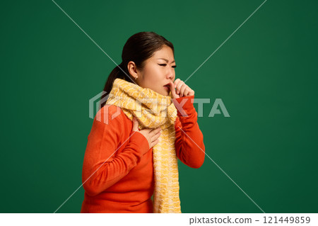 Woman coughing into hand, wearing scarf, showing symptoms of viral respiratory illness, bronchitis, or flu causing persistent cough. 121449859