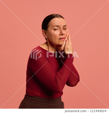 Elderly woman in red presses her cheek with both hands, showing toothache or facial pain, against soft pink background. Elderly woman in red presses her cheek with both hands, showing toothache or facial pain, against soft pink background. 121449924