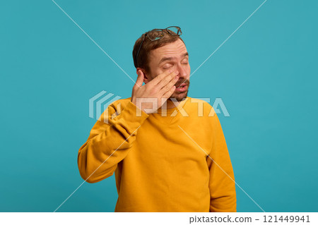 Man in yellow sweater rubbing eyes with glasses resting on head and tired expression against blue studio background. Man in yellow sweater rubbing eyes with glasses resting on head and tired expression against blue studio background. 121449941