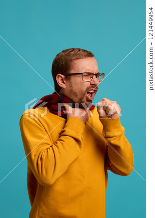 Portrait of young man in glasses suffering from sore throat and cough, warming neck with scarf against blue studio background. 121449954