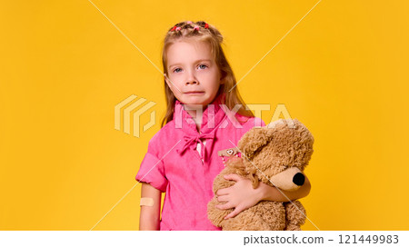 Girl in pink dress tearfully glancing sideways, clutching teddy bear for comfort after vaccination against yellow studio background. Girl in pink dress tearfully glancing sideways, clutching teddy bear for comfort after vaccination against yellow studio background. 121449983