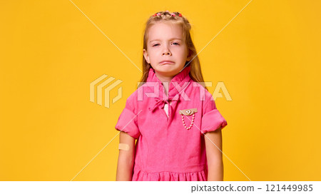 Girl in pink dress tearfully looking at camera with patch on hand after painful vaccination against yellow studio background. Girl in pink dress tearfully looking at camera with patch on hand after painful vaccination against yellow studio background. 121449985