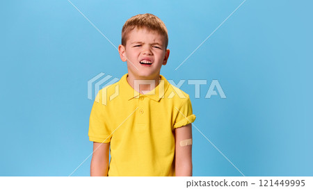 Boy grimacing slightly, bandaged arm visible, creating expressive moment of exaggerated discomfort against blue studio background. 121449995