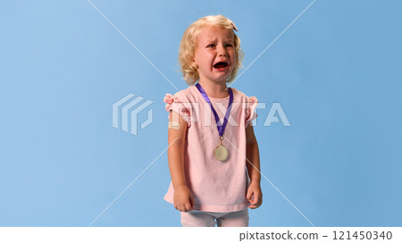 Upset, crying child, little girl in pink outfit with patch after vaccination on hand, wearing gold medal around neck against blue studio background. Upset, crying child, little girl in pink outfit with patch after vaccination on hand, wearing gold medal around neck against blue studio background. 121450340
