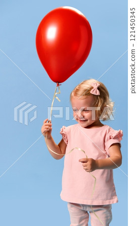 Little charming child, girl in pink clothes, smiling softly while holding red balloon against blue studio background. Little charming child, girl in pink clothes, smiling softly while holding red balloon against blue studio background. 121450345