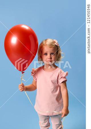Little blonde child in pink outfit with bandage on hand after vaccination holding red balloon with sad expression against blue studio background. Little blonde child in pink outfit with bandage on hand after vaccination holding red balloon with sad expression against blue studio background. 121450346