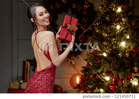 Playful Woman Holding Gift by Christmas Tree. Playful Woman Holding Gift by Christmas Tree. 121450711
