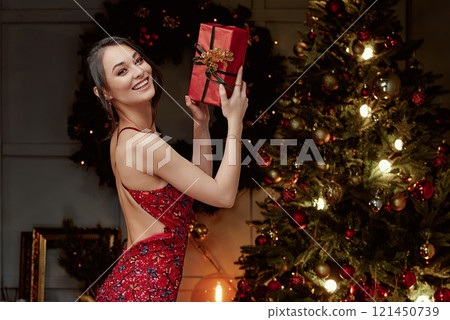 Happy Woman Holding Christmas Gift Next to Festive Tree. 121450739
