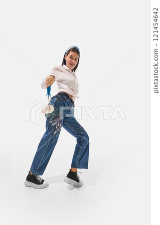 Painter in white shirt and colorful jeans leaning backward, raising paint roller playfully with joyful expression against white studio background. 121454642