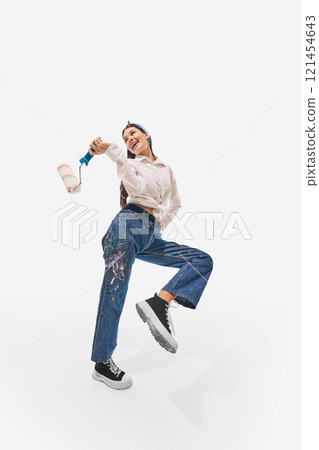 Painter wearing white shirt and paint-splattered jeans, holding paint roller, smiling vibrantly while posing against white studio background. Painter wearing white shirt and paint-splattered jeans, holding paint roller, smiling vibrantly while posing against white studio background. 121454643