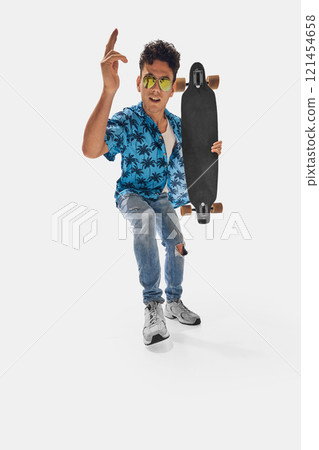 Wide angle photo of skateboarder wearing tropical blue shirt and ripped jeans, pointing upwards while holding skateboard confidently 121454658